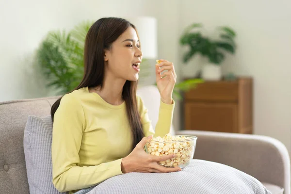 年轻的亚洲女人把一碗爆米花放在枕头上 一边吃爆米花 一边坐在舒适的大沙发上 在家里的客厅里看电视 — 图库照片