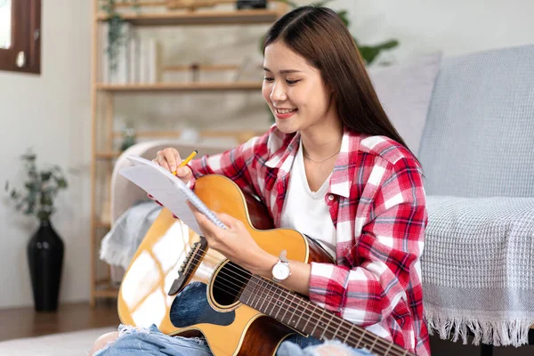 Young woman is compose song and writing lyrics on notebook while putting guitar on the legs and sitting on the floor in living room at home.