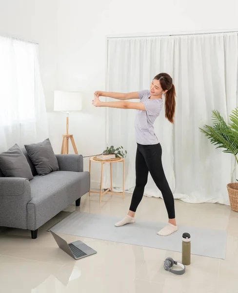 Young woman is taking yoga lesson online on laptop and workout yoga exercise while standing to stretching arms on mat in her living room at home.