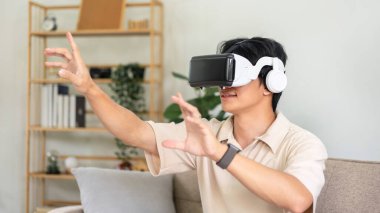 Sanal gerçeklik gözlüğü takan genç Asyalı bir adam sanal gerçeklik deneyimi olan oyunlar oynarken parmaklarıyla havaya dokunuyor ve oturma odasında kanepede oturuyor..