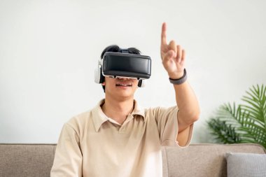 Sanal gerçeklik gözlüğü takan genç Asyalı adam video izlerken ve sanal deneyimle oyunlar oynarken işaret parmaklarıyla havaya dokunurken ve oturma odasında kanepede otururken.