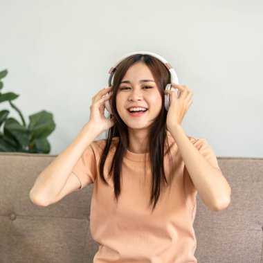 Genç Asyalı kadın iki eliyle kulaklığa dokunarak müzik dinliyor ve rahat bir kanepede oturup evdeki oturma odasında dinlenirken şarkıyı söylüyor..