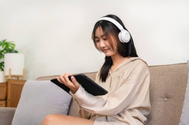 Kadın girişimci büyük rahat bir koltukta oturuyor ve evde dinlenirken film izlemek ve müzik dinlemek için tablet tutarken kulaklık takıyor..