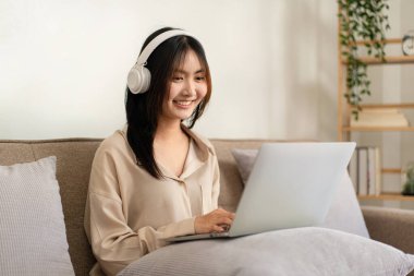 Kadın girişimci, evdeki oturma odasında sohbet ve video konferans için kulaklık takarken büyük rahat bir kanepeye oturmuş yastığa dizüstü bilgisayar koyuyor..