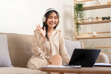 Kadın girişimci büyük rahat bir koltukta oturuyor ve evde oturma odasındaki video konferansında buluşmak ve karşılamak için tablet kullanırken kulaklık takıyor..