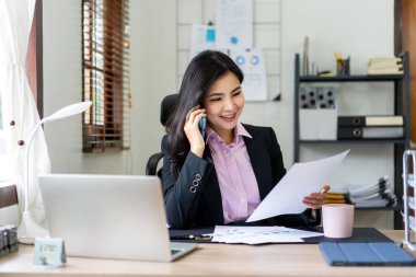 Takım elbiseli genç iş kadını, akıllı telefon ve iş belgelerini okurken aynı zamanda stratejinin analizi ve ofiste yeni iş planları üzerinde çalışıyor..