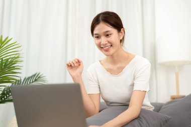 Genç Asyalı bir kadın evdeki oturma odasında rahat koltukta otururken ve çalışırken günlük giysiler giyerken dizüstü bilgisayarında proje ve iş verileri hakkında yeni fikirler yazıyor..