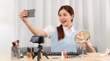 Genç güzellik uzmanı, sosyal medyada ya da web blogcusu stüdyoda paylaşmak üzere kameraya video kaydederken güzellik ekipmanları ve makyaj aksesuarlarıyla selfie çekmek için akıllı telefon kullanıyor..