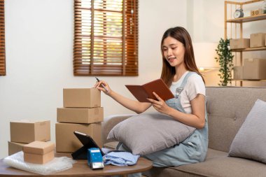 Genç girişimci, müşteri koltuğunda rahat bir şekilde otururken ve ofisteki oturma odasında çalışırken karton kutularda müşteri verilerini okuyor ve müşterilere ev teslimatı için adres yazıyor..