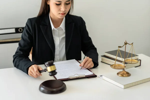 Business lawyer woman holding justice hammer and reading business contract to writing information on document while working on the table with brass scales in law firm office.