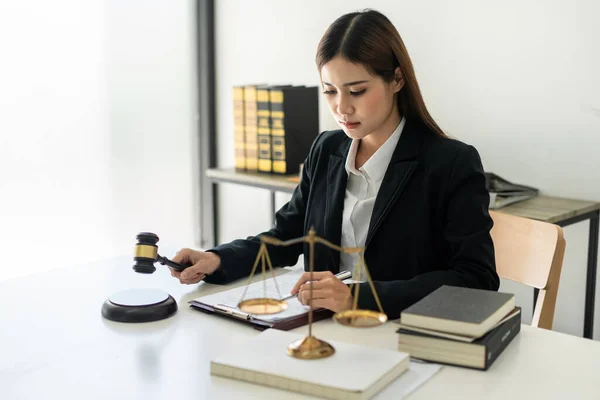Business lawyer woman holding justice hammer and reading business contract to writing information on document while working on the table with brass scales in law firm office.
