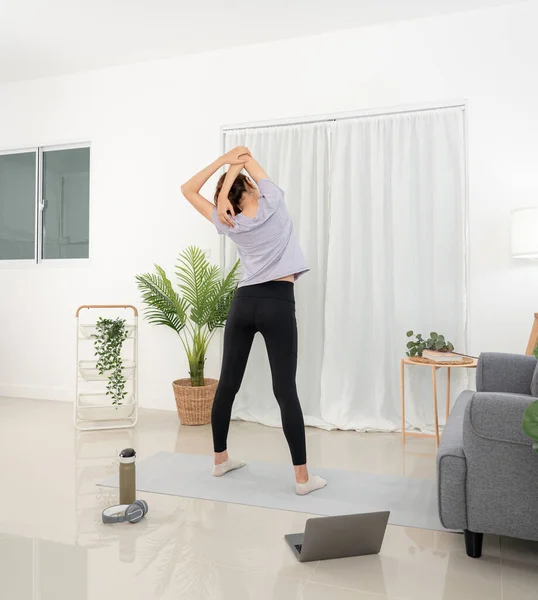 stock image Young woman is watching yoga lessons online on laptop and stretching upper arms before workout yoga exercise on mat in living room.