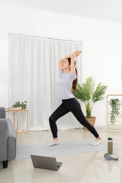 Young woman is taking yoga lesson online on laptop and standing to practicing yoga with pose warrior one exercise while workout on mat in her living room at home.
