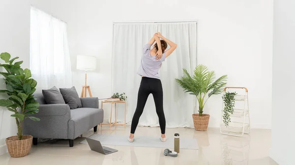 Young woman is watching yoga lessons online on laptop and stretching upper arms before workout yoga exercise on mat in living room.