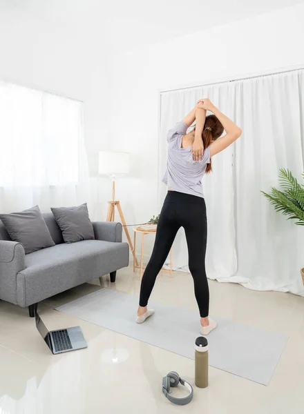 Young woman is watching yoga lessons online on laptop and stretching upper arms before workout yoga exercise on mat in living room.