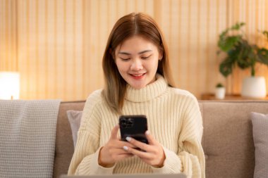 Genç Asyalı kadın akıllı telefondan iş verilerine bakıyor ve süveter giyerken dizüstü bilgisayarda çalışıyor ve tatilde evdeki Noel dekorasyonunun rahat oturma odasındaki kanepede çalışıyor..