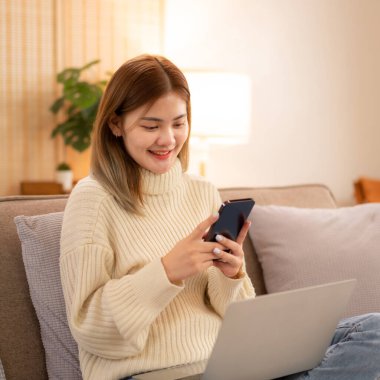 Genç Asyalı kadın akıllı telefondan iş verilerine bakıyor ve süveter giyerken dizüstü bilgisayarda çalışıyor ve tatilde evdeki Noel dekorasyonunun rahat oturma odasındaki kanepede çalışıyor..