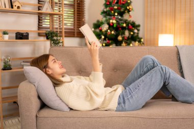 Genç Asyalı kadın süveter giyer ve kitap okurken tatilde dinlenmek için koltukta uzanır evinde Noel süslemelerinin rahat oturma odasında.