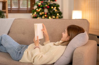 Genç Asyalı kadın süveter giyer ve kitap okurken tatilde dinlenmek için koltukta uzanır evinde Noel süslemelerinin rahat oturma odasında.