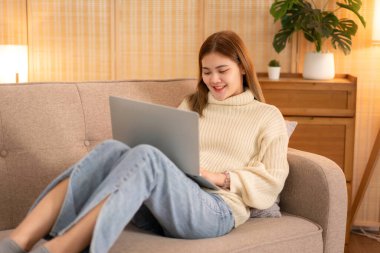 Genç Asyalı kadın süveter giyer ve iş verilerini dizüstü bilgisayarda yazarken evdeki Noel dekorasyonunun rahat oturma odasında çalışmak için kanepeye yaslanır..