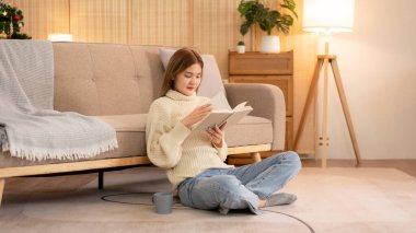 Genç Asyalı kadın, kahve içip kitap okurken süveter giyer ve tatilde dinlenmek için evde Noel süslemelerinin olduğu sıcak oturma odasında yere oturur..