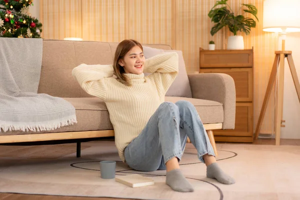 年轻的亚洲女人穿着毛衣 双手交叉在脑后 坐在舒适舒适的圣诞装饰客厅的地板上放松自己 — 图库照片
