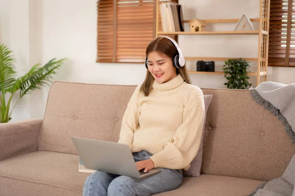 Young asian woman chatting and meeting in video conference with partner on laptop while wearing headphone and sitting to working in holiday on sofa in cozy living room of christmas decoration at home.