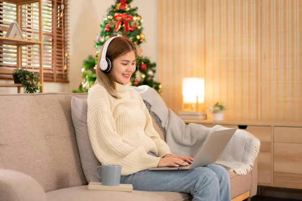 Young asian woman chatting and meeting in video conference with partner on laptop while wearing headphone and sitting to working in holiday on sofa in cozy living room of christmas decoration at home.