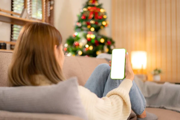 Young woman meeting in video conference with colleague on blank screen smartphone while wearing sweater and lying to working in holiday on sofa in cozy living room of christmas decoration