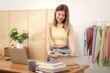 Kadın tasarımcı girişimci dizüstü bilgisayardan siparişleri kontrol ediyor ve giysi ürünlerini karton kutuya koyup eve nakliye için hazırlanıyor..