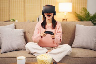 Evde oturma odasında dinlenirken elinde joystick ile sanal gerçeklikle video oyunu oynayan süveter ve sanal gözlük takan genç bir lezbiyen kadın..