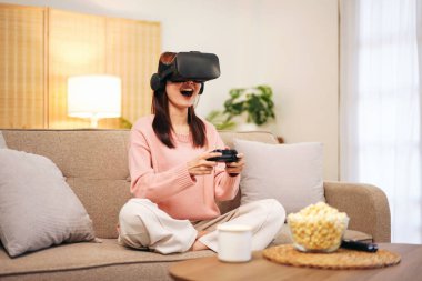 Evde oturma odasında dinlenirken elinde joystick ile sanal gerçeklikle video oyunu oynayan süveter ve sanal gözlük takan genç bir lezbiyen kadın..