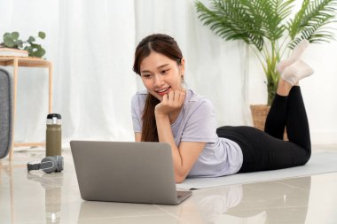 Genç bir kadın yoga minderinde uzanıyor, çenesi dinlenmek ve dizüstü bilgisayarda film izlemek için dinlenirken oturma odasında yoga egzersizine ara veriyor..