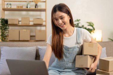 Genç girişimci online siparişleri kontrol etmek için dizüstü bilgisayar kullanıyor ve kanepede rahat bir şekilde otururken karton kutular tutuyor ve ev ofisinin oturma odasında çalışıyor..