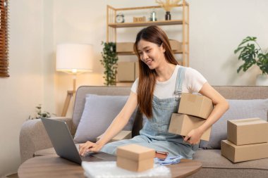 Genç girişimci online siparişleri kontrol etmek için dizüstü bilgisayar kullanıyor ve kanepede rahat bir şekilde otururken karton kutular tutuyor ve ev ofisinin oturma odasında çalışıyor..