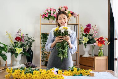 Çiçekçi dükkanında çiçek buketi hazırlarken ve dizayn ederken elinde kasımpatı tutarken gülümseyen ve eğlenen önlüklü kadın..