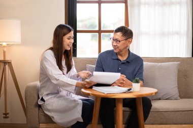 Kadın doktor erkek yaşlı hastayı evinde ziyaret ediyor ve sağlık kontrolünün detaylarını açıklayan belgeye işaret ediyor..