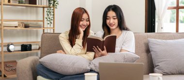 Genç lezbiyen çift evde oturma odasındaki büyük rahat kanepede oturup dinlenirken birlikte kitap okuyorlar..