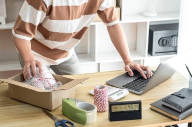 Erkek girişimci çevrimiçi siparişleri kontrol etmek için dizüstü bilgisayar kullanıyor. Ürünü karton kutulara dolduruyor. Müşteri göndermeye hazırlanıyor..