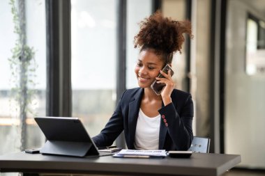 Takım elbiseli Afrikalı Amerikalı iş kadını, tabletle ilgili muhasebe bilgilerini okuyor ve dijital tablet üzerinde çalışırken akıllı telefonla konuşuyor..