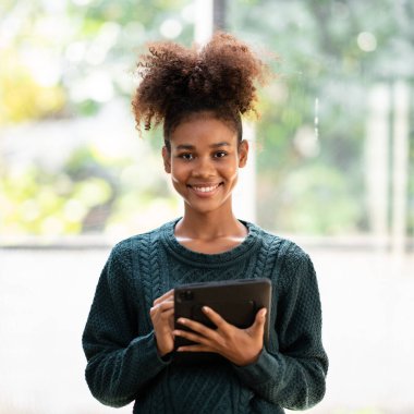 Kazak giyen Afrikalı Amerikalı bir kadın üniversitede dururken online eğitim ve bilgi dersi aldıktan sonra dijital tablet hakkında bilgi ve ödev yazıyor..