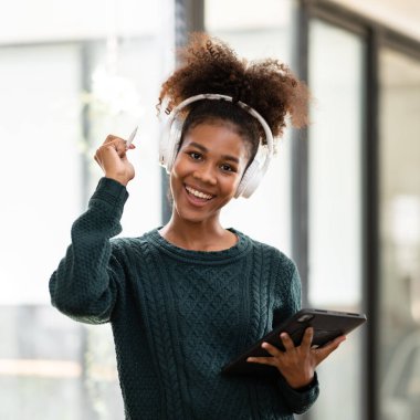 Üniversitede eğitim ve bilgi edinirken online ders çalışmak ve dijital tablet hakkında bilgi yazmak için kazak ve kulaklık takan Afrikalı Amerikalı bir kadın öğrenci..