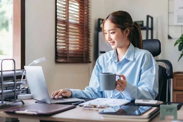 Businesswoman is thoughtful about accounting of business and drinking hot chocolate while using laptop to working and typing information of new business project in modern office.