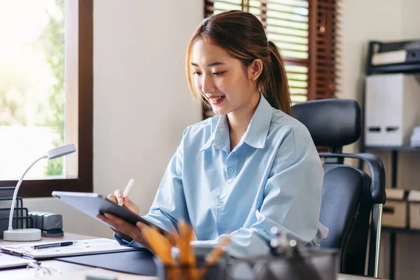 Businesswoman is reading information in document and taking notes in tablet while thoughtful about accounting of business and working about new business project at modern office.
