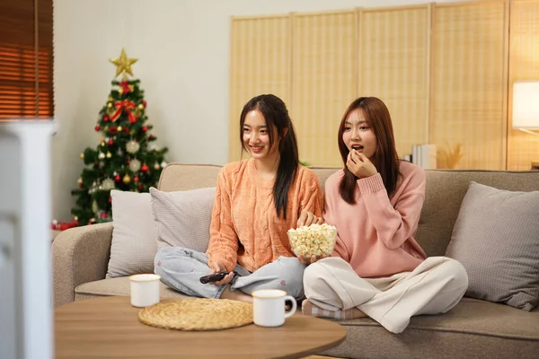 Young woman lesbian couple in sweater watching cinema with excited face and holding tv remote control to switching cinema channel while eating delicious popcorn and spending time together.