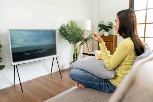 Young asian woman is watching movie on television and holding remote control to switching channel while eating popcorn and sitting on the big comfortable sofa in living room at home.