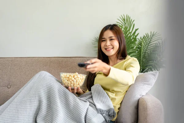 年轻的亚洲女人在电视上看电影 一边拿着爆米花 一边手握遥控装置 一边打开频道 躺在家里客厅里舒适的大沙发上 — 图库照片