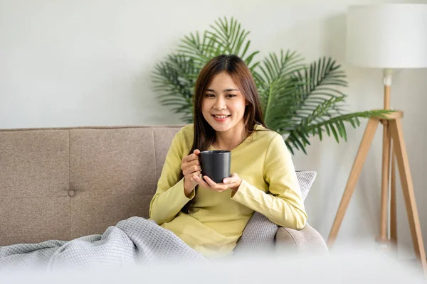 年轻的亚洲女人正在喝咖啡 躺在舒适的大沙发上 在家客厅里看电视上的电影 — 图库照片