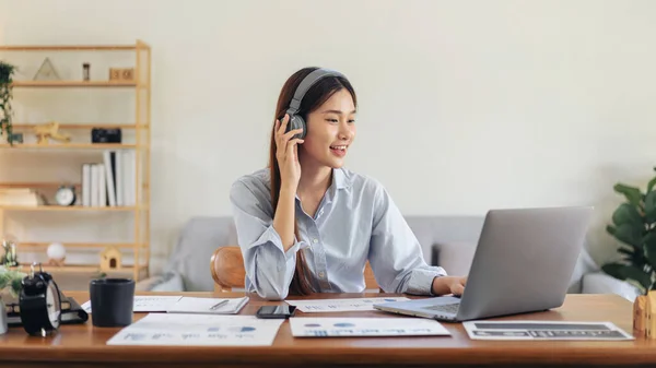 Female freelance is using laptop and wearing headphone to meeting video conference with colleague while working on the table in living room at home office.
