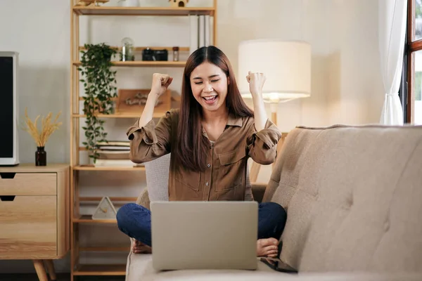 女性起業家はラップトップで新しいビジネスプロジェクトを読んで 自宅のオフィスのリビングルームで大きな快適なソファに座っている間に正常に仕事を祝うために拳を上げる — ストック写真